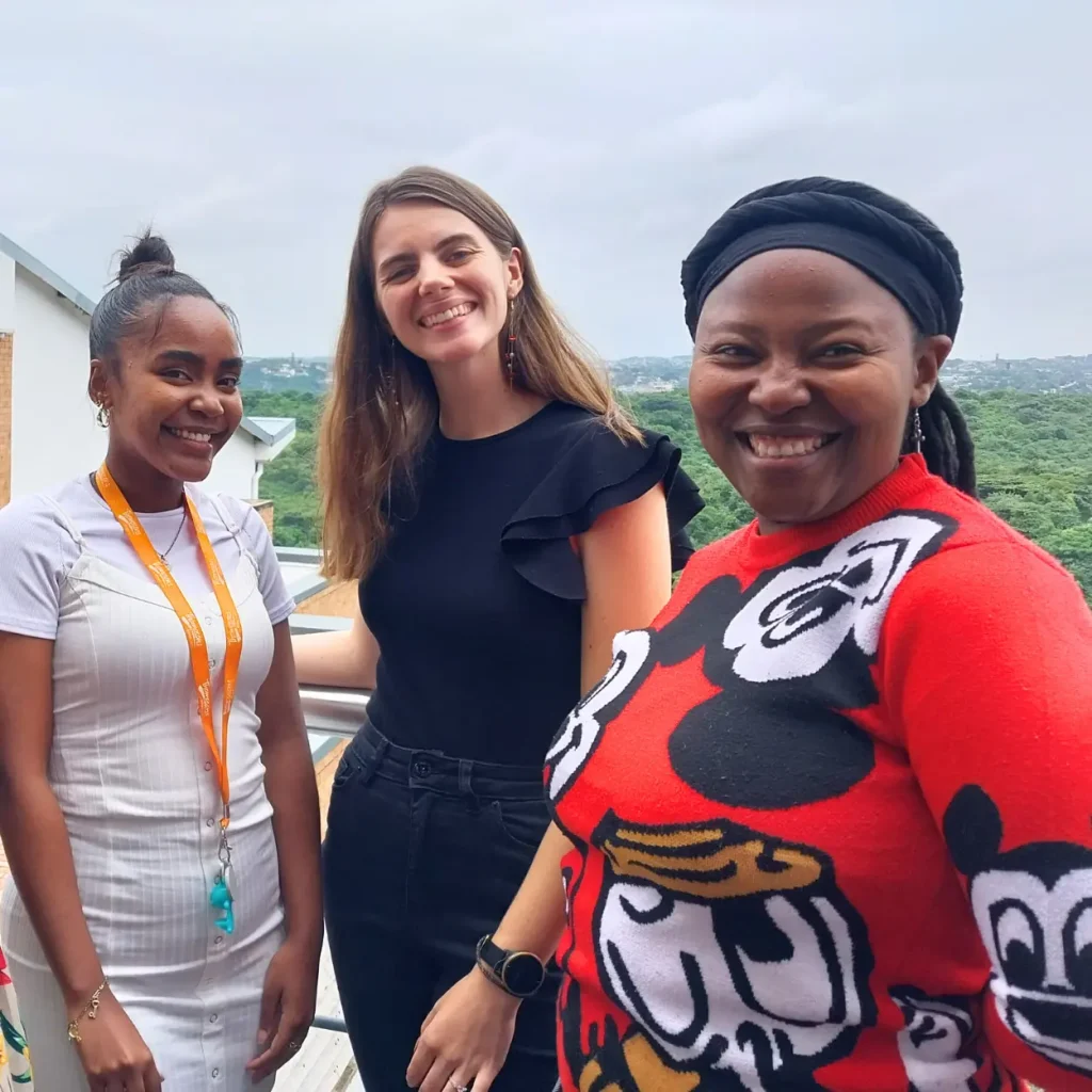 Three smiling women standing on a balcony with a scenic green landscape in the background.